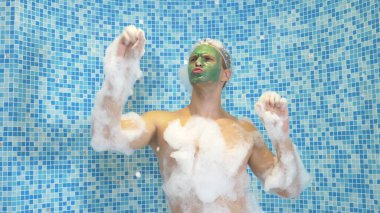 Yüzünde yeşil bir maske ve duş bonesi olan genç bir adam köpükle banyo yapıyor. Adam banyoda dans ediyor, vücudu kalın beyaz köpükle kaplı. Boşluğu kopyala