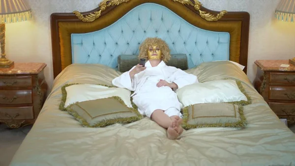 Wanita pirang muda dengan topeng emas di wajahnya menonton televisi berbaring di tempat tidur emas mewah . — Stok Foto