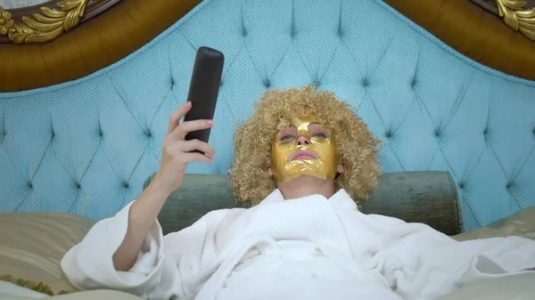 Jonge blonde vrouw met een gouden masker op haar gezicht tv kijken liggend op een luxe gouden bed. — Stockfoto