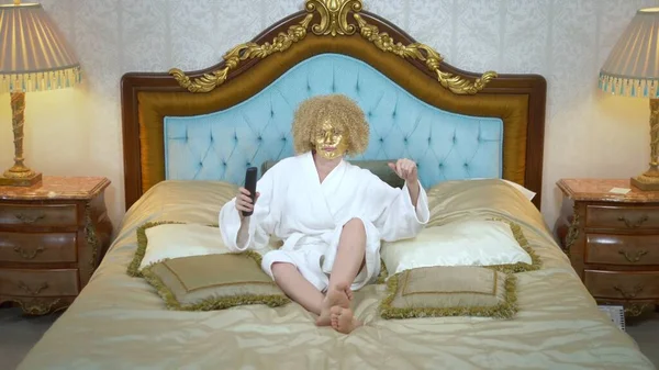 Junge blonde Frau mit goldener Maske im Gesicht vor dem Fernseher auf einem luxuriösen goldenen Bett liegend. — Stockfoto