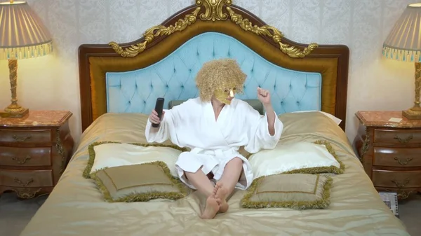 Jovem loira com uma máscara dourada em seu rosto assistindo televisão deitada em uma luxuosa cama dourada . — Fotografia de Stock