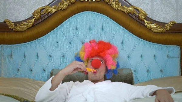 A young man in a white coat and a clown wig lies on a luxurious golden bed and eats a lollipop on a stick. concept of humor, adventure of strange people. — Stockfoto