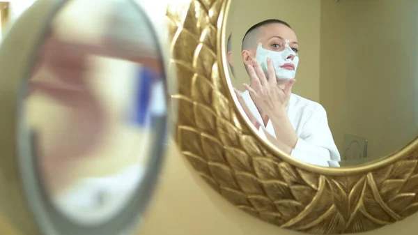 Chica calva en un abrigo blanco pone una máscara de belleza en su cara, mirando en el espejo en el baño . — Foto de Stock