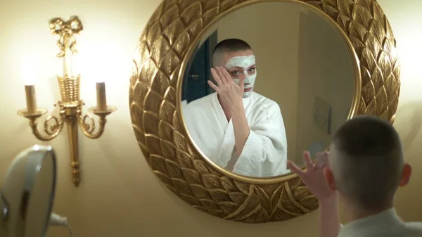 Holohlavá dívka v bílém plášti si nasadí masku krásy na obličej, dívá se do zrcadla v koupelně. — Stock fotografie