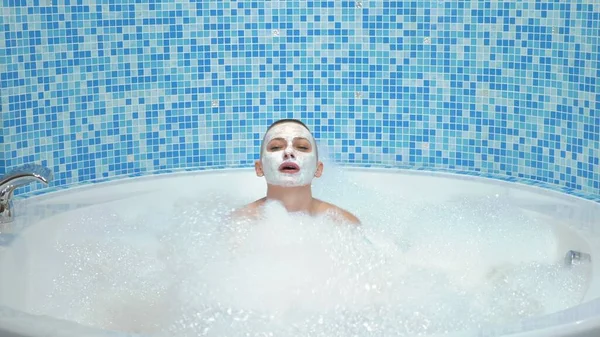 Uma jovem careca com uma máscara no rosto encontra-se em um banho com espuma, ela sorri e sopra na espuma — Fotografia de Stock