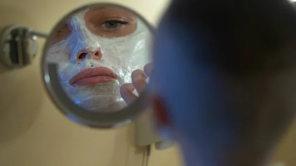 Ragazza calva in un cappotto bianco mette una maschera di bellezza sul suo viso, guardando nello specchio in bagno . — Foto Stock