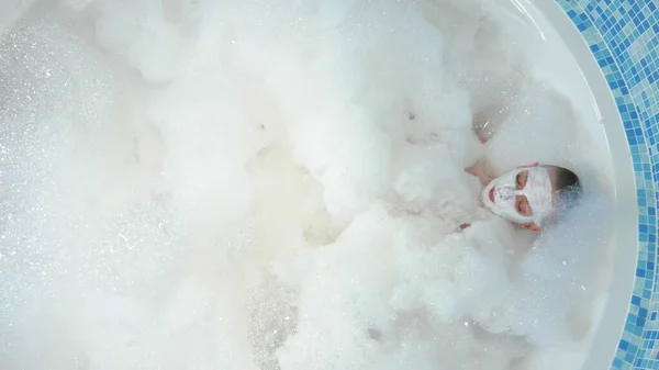 Utsikt uppifrån. En attraktiv ung skallig kvinna med mask i ansiktet ligger i ett badkar med tjockt frodigt skum. vila, avkoppling — Stockfoto