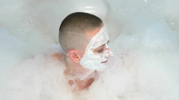 A bald young woman with a mask on her face lies in a bath with foam, she smiles and blows on the foam — Stock Photo, Image