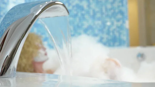 In focus water pouring from a faucet into a bath, on a blurred background Young woman in a face mask take a bubble bath — 스톡 사진