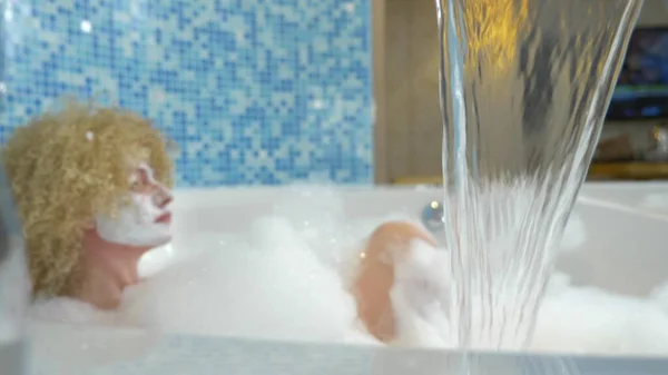 Im Fokus Wasser, das aus einem Wasserhahn in eine Badewanne fließt, vor verschwommenem Hintergrund nimmt eine junge Frau mit Gesichtsmaske ein Schaumbad — Stockfoto