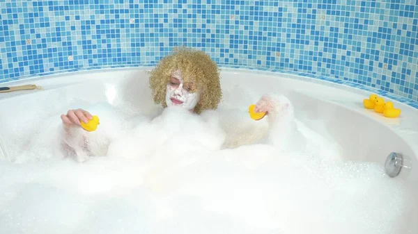 Schöne junge blonde Frau mit einer Maske auf dem Gesicht beim Baden mit Blasen spielt mit einer gelben Ente. Humorvolles Konzept — Stockfoto