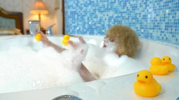 Belle jeune femme blonde avec un masque sur le visage tout en prenant un bain avec des bulles joue avec un canard jaune. concept humoristique — Photo