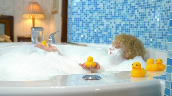 Mulher loira jovem bonita com uma máscara em seu rosto enquanto toma um banho com bolhas está brincando com um pato amarelo. conceito humorístico — Fotografia de Stock