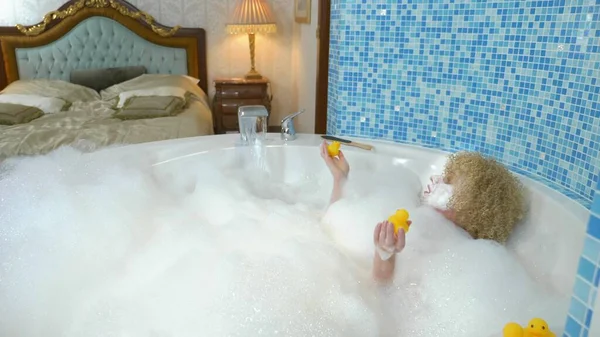 Mulher loira jovem bonita com uma máscara em seu rosto enquanto toma um banho com bolhas está brincando com um pato amarelo. conceito humorístico — Fotografia de Stock