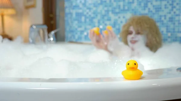 Beautiful young blond woman with a mask on her face while taking a bath with bubbles is playing with a yellow duck. humorous concept — 스톡 사진