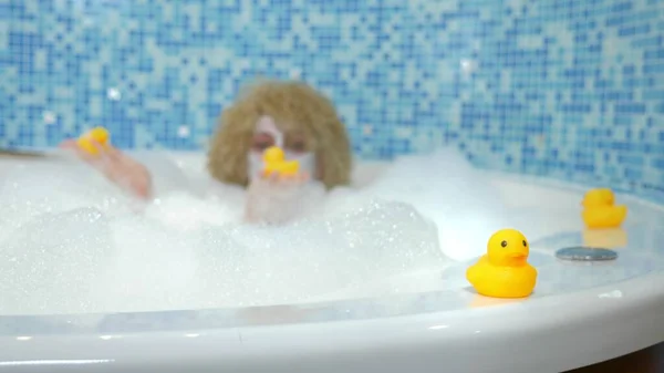 Beautiful young blond woman with a mask on her face while taking a bath with bubbles is playing with a yellow duck. humorous concept — 스톡 사진