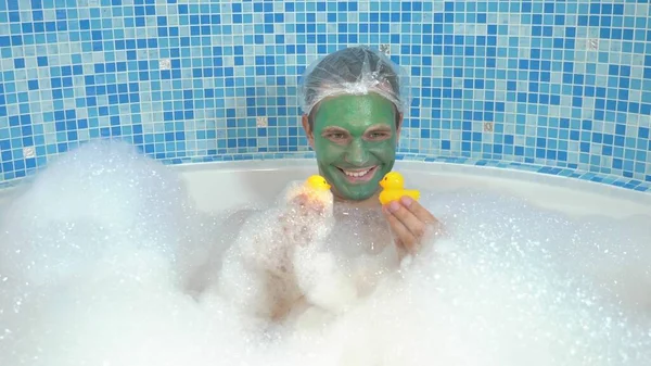 Een jonge man met een groen cosmetisch masker op zijn gezicht en in een douchemuts neemt een bad met schuim en speelt met een gele speelgoedeend. Humorvol concept, avonturen van vreemde mensen. — Stockfoto