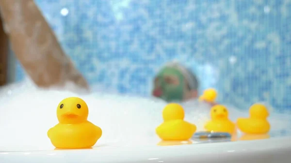 Um jovem com uma máscara cosmética verde em seu rosto e em uma touca de banho toma um banho com espuma e brinca com um pato de brinquedo amarelo. Conceito humorístico, aventuras de pessoas estranhas . — Fotografia de Stock