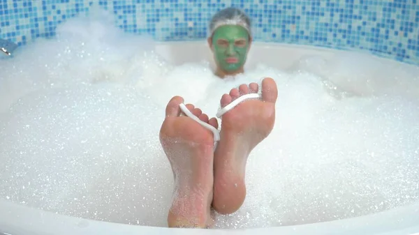 A young man in a shower cap and with a green cosmetic mask on his face lies in the bathroom with copious foam. his feet with finger dividers are visible from the water, in focus — 스톡 사진