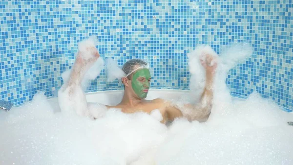 Um jovem com uma máscara verde em seu rosto e em uma touca de banho toma um banho com espuma. o cara está dançando na casa de banho, seu corpo está coberto com espessa espuma branca. espaço de cópia — Fotografia de Stock