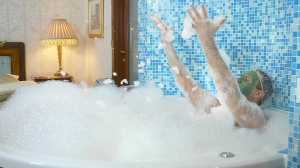 A young man with a green mask on his face and in a shower cap takes a bath with foam. the guy is dancing in the bathroom, his body is covered with thick white foam. copy space — 스톡 사진