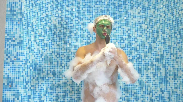 Un giovane uomo con una maschera verde sul viso e con una cuffia da doccia si fa un bagno con schiuma. il ragazzo sta ballando in bagno, il suo corpo è coperto di spessa schiuma bianca. copia spazio — Foto Stock