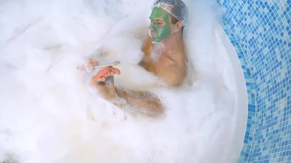Ein Mann mit Duschkappe und grüner Gesichtsmaske liegt in einem Bad mit reichlich Schaum und wäscht sich den Fuß mit Bimsstein. Hygiene mit Peeling im Bad. Blick von oben — Stockfoto