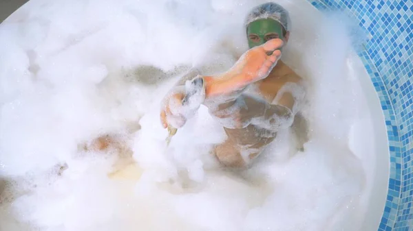 Ein Mann mit Duschkappe und grüner Gesichtsmaske liegt in einem Bad mit reichlich Schaum und wäscht sich den Fuß mit Bimsstein. Hygiene mit Peeling im Bad. Blick von oben — Stockfoto