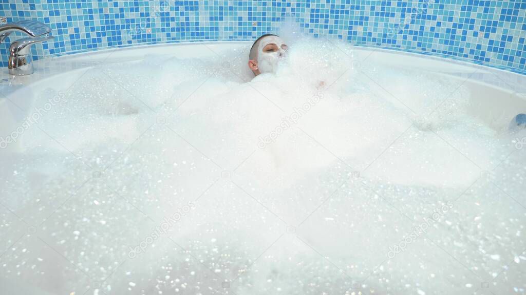 a bald young woman with a mask on her face lies in a bath with foam, she smiles and blows on the foam