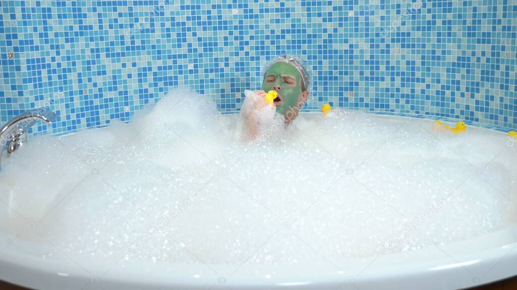 a young man with a green cosmetic mask on his face and in a shower cap takes a bath with foam and plays with a yellow toy duck. Humorous concept, adventures of strange people.