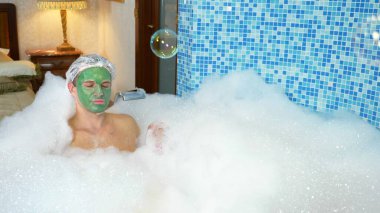 Banyoda bol köpüklü bir şekilde banyo küvetinde uzanan banyo bonesi takmış, kilden maskeli duygusal yakışıklı adam sabun köpükleri üflüyor. Boşluğu kopyala