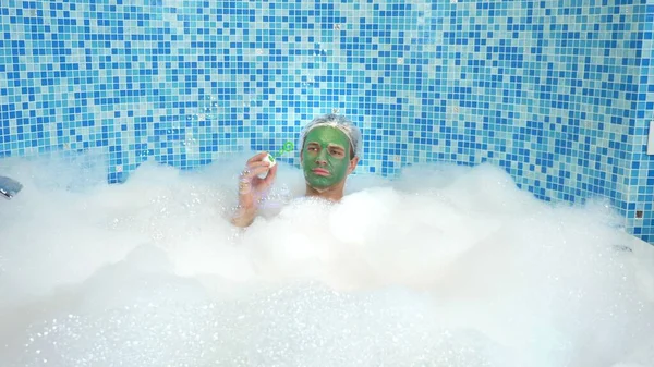 Emotional cute man with clay mask in a bathing cap, blowing soap bubbles while lying in a bathtub with plentiful foam. humorous concept. copy space — Stock Photo, Image