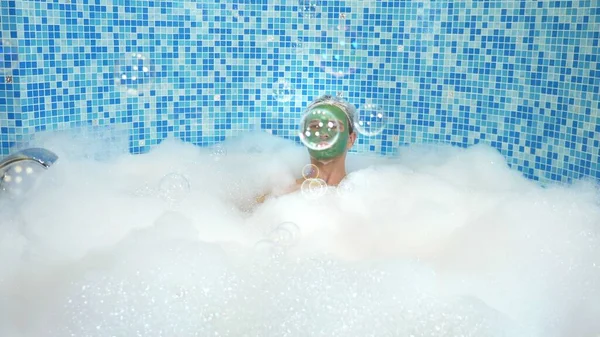 Emotional cute man with clay mask in a bathing cap, blowing soap bubbles while lying in a bathtub with plentiful foam. humorous concept. copy space — 스톡 사진