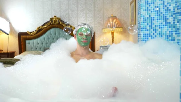 Homem bonito emocional com máscara de barro em uma touca de banho, soprando bolhas de sabão enquanto deitado em uma banheira com espuma abundante. conceito humorístico. espaço de cópia — Fotografia de Stock