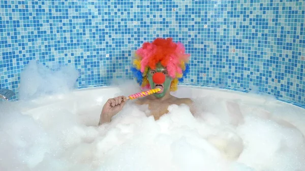 Aranyos bohóc egy fürdőkádban bőséges habbal megeszik egy nyalókát egy boton. humoros fogalom. — Stock Fotó