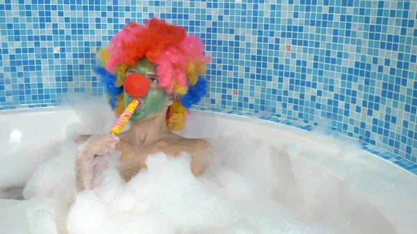 泡の多い浴槽で可愛いピエロさんが棒にロリポップを食べます。ユーモアのあるコンセプト. — ストック写真