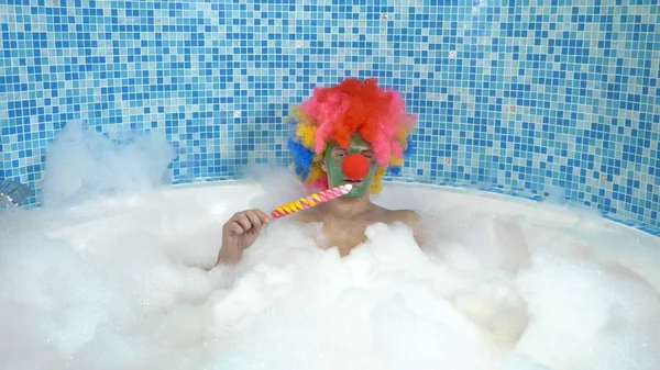 Słodki klaun w wannie z dużą ilością pianki zjada lizaka na patyku. humorystyczna koncepcja. — Zdjęcie stockowe