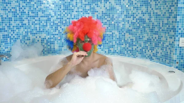 Der süße Clown in einer Badewanne mit reichlich Schaum frisst einen Lutscher auf einem Stock. Humorvolles Konzept. — Stockfoto