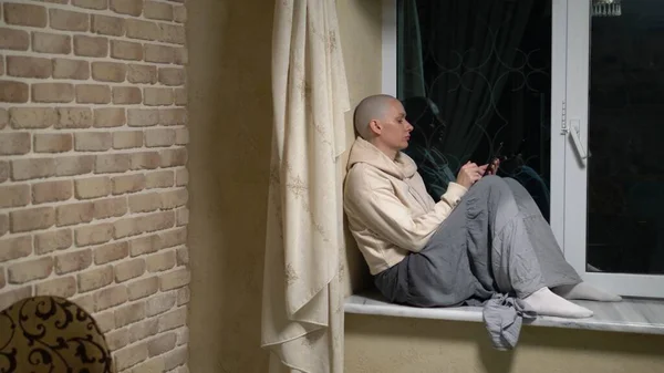 Mujer calva triste se sienta en un alféizar de la ventana por la noche y utiliza el teléfono — Foto de Stock