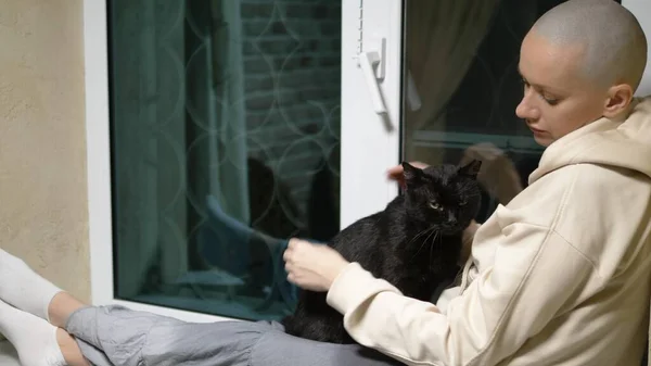 Mulher careca triste senta-se à noite em um peitoril da janela pela janela e acaricia um gato preto — Fotografia de Stock