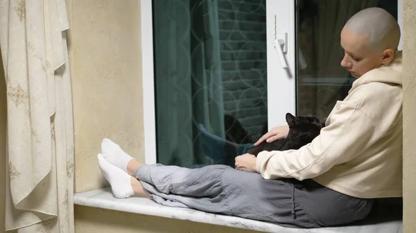 Üzgün, kel bir kadın gece pencerenin kenarında oturur ve siyah bir kediyi okşar. — Stok fotoğraf