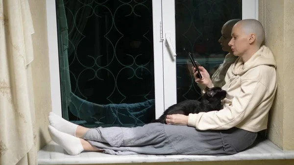 Грустная лысая женщина сидит ночью на подоконнике у окна, гладит черную кошку и пользуется смартфоном — стоковое фото