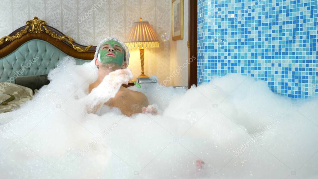 emotional cute man with clay mask in a bathing cap blowing soap bubbles lying in a bathtub with plentiful foam in a luxurious bathroom. copy space