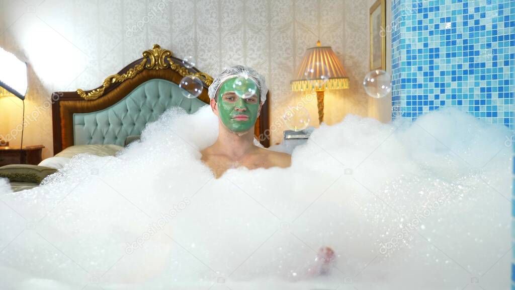 emotional cute man with clay mask in a bathing cap, blowing soap bubbles while lying in a bathtub with plentiful foam. humorous concept. copy space