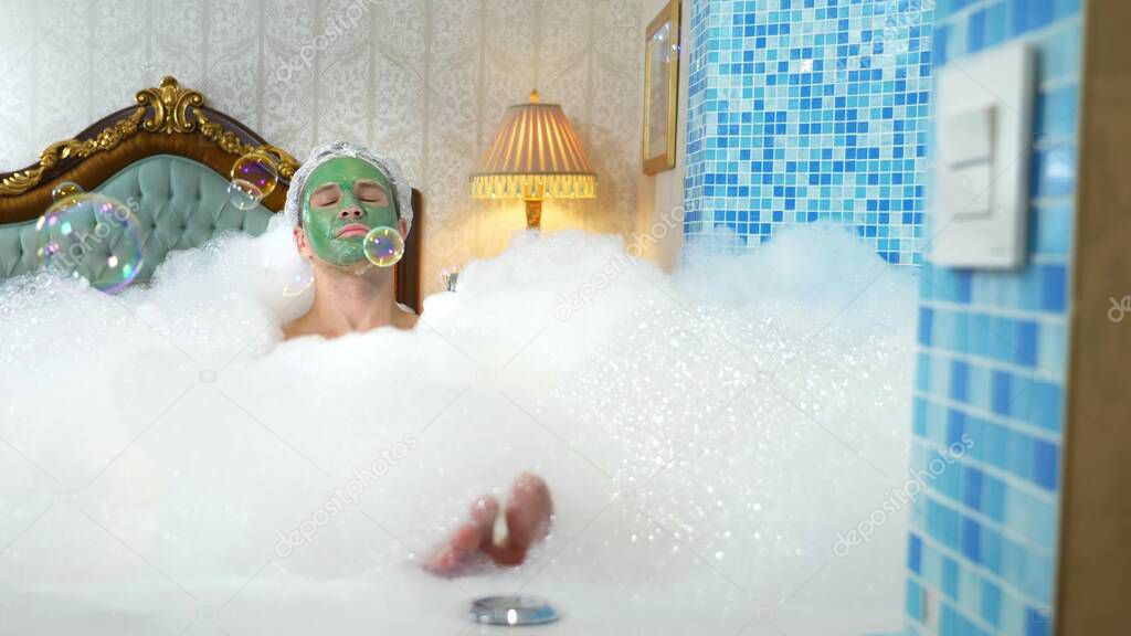 emotional cute man with clay mask in a bathing cap blowing soap bubbles lying in a bathtub with plentiful foam in a luxurious bathroom. copy space
