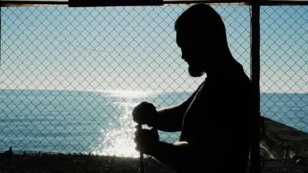 剪影。 战斗前,拳击手用弹性绷带包扎他的手. 在海滩上的体育馆里，在蓝色的海面上 — 图库视频影像