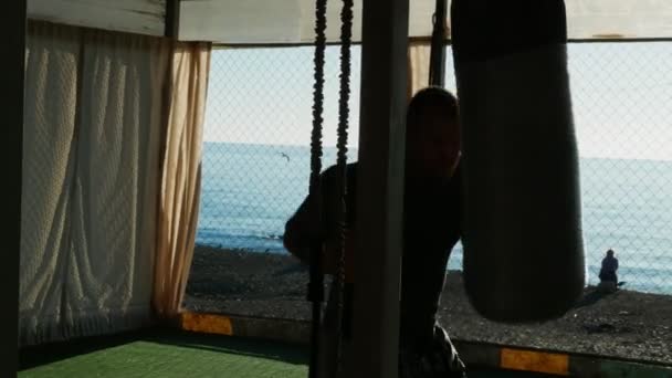 Silhouette. Boxer beats punching bag in open gym on the beach with sea view. — Stock Video