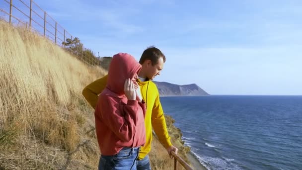 Par, man och kvinna i färgglada hoodies står på en klippa beundra utsikten över havet — Stockvideo