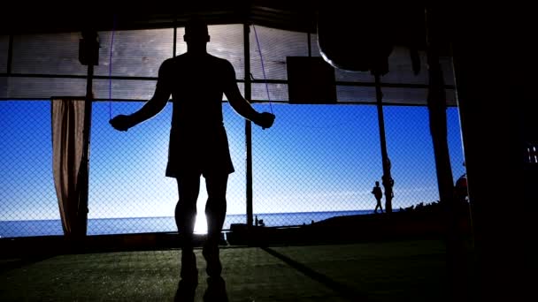 Primer plano. Super cámara lenta. silueta. hombre deportivo saltar la cuerda en el gimnasio al aire libre en la playa con vistas al mar azul y cielo despejado — Vídeos de Stock