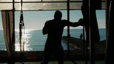 Silüet. Boksör, açık salonda deniz manzaralı kum torbasını yener..